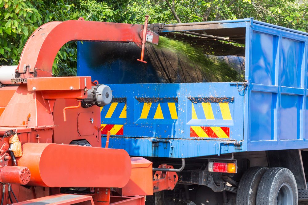 Wood chipper blowing green debris into blue dump truck during a debris removal job